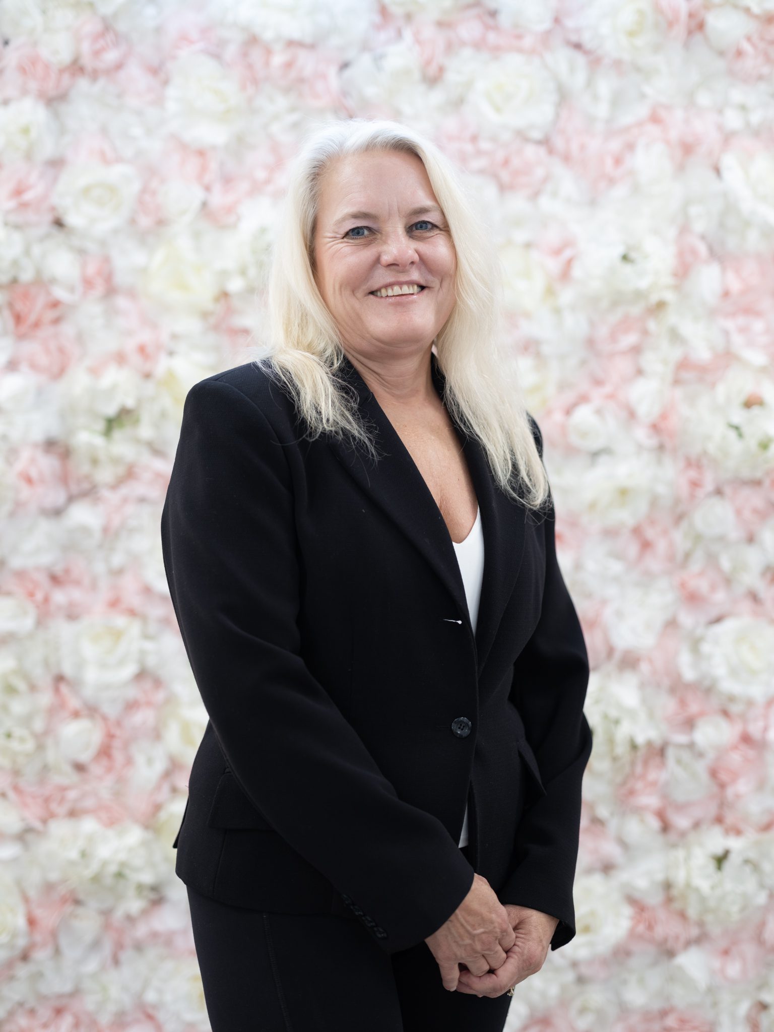 Woman in black blazer smiling against a floral backdrop.