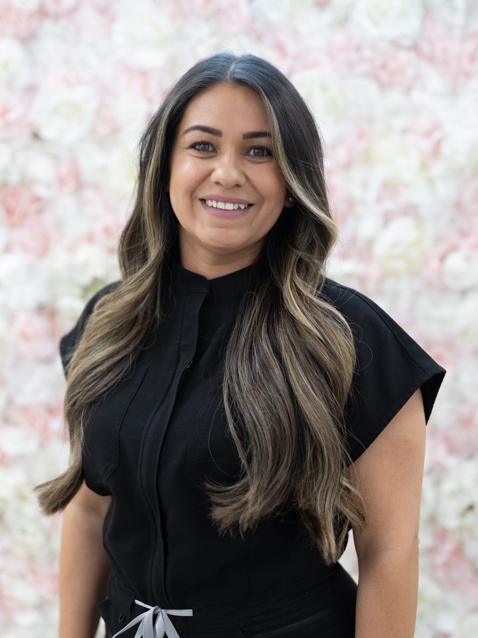 Smiling woman with long wavy hair in black top against floral background.