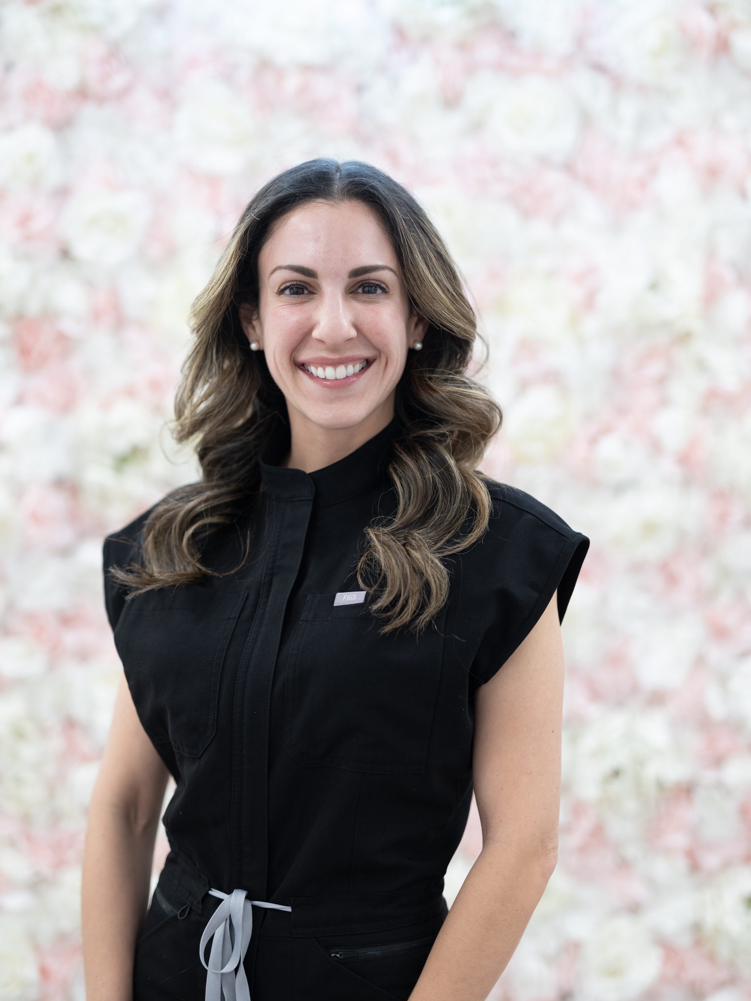 Smiling woman with wavy hair in a black sleeveless top against a floral background.
