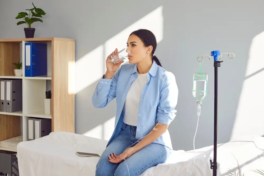 Woman with IV line drinking water on bed